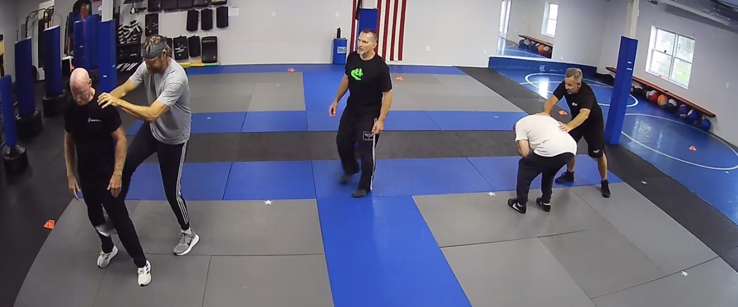 Partners working a self-defense drill on the mat while an instructor supervises